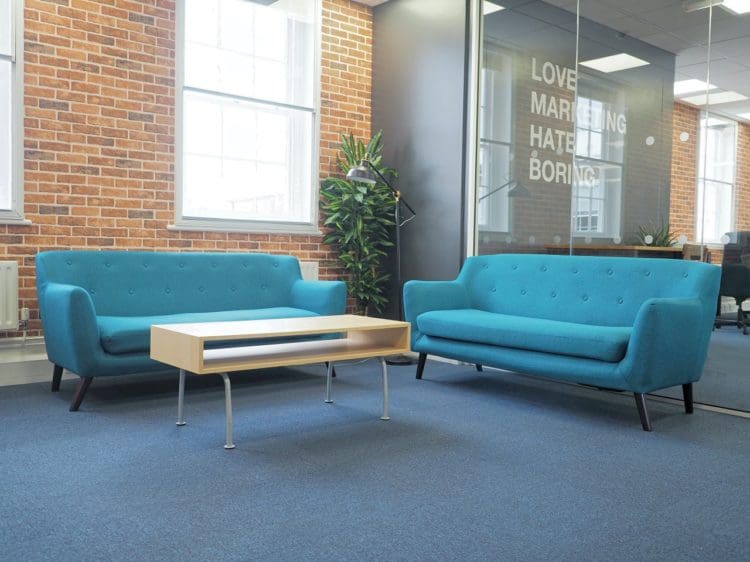An informal breakout space with bright blue sofa and a wood coffee table to contrast the brick effect wallpaper